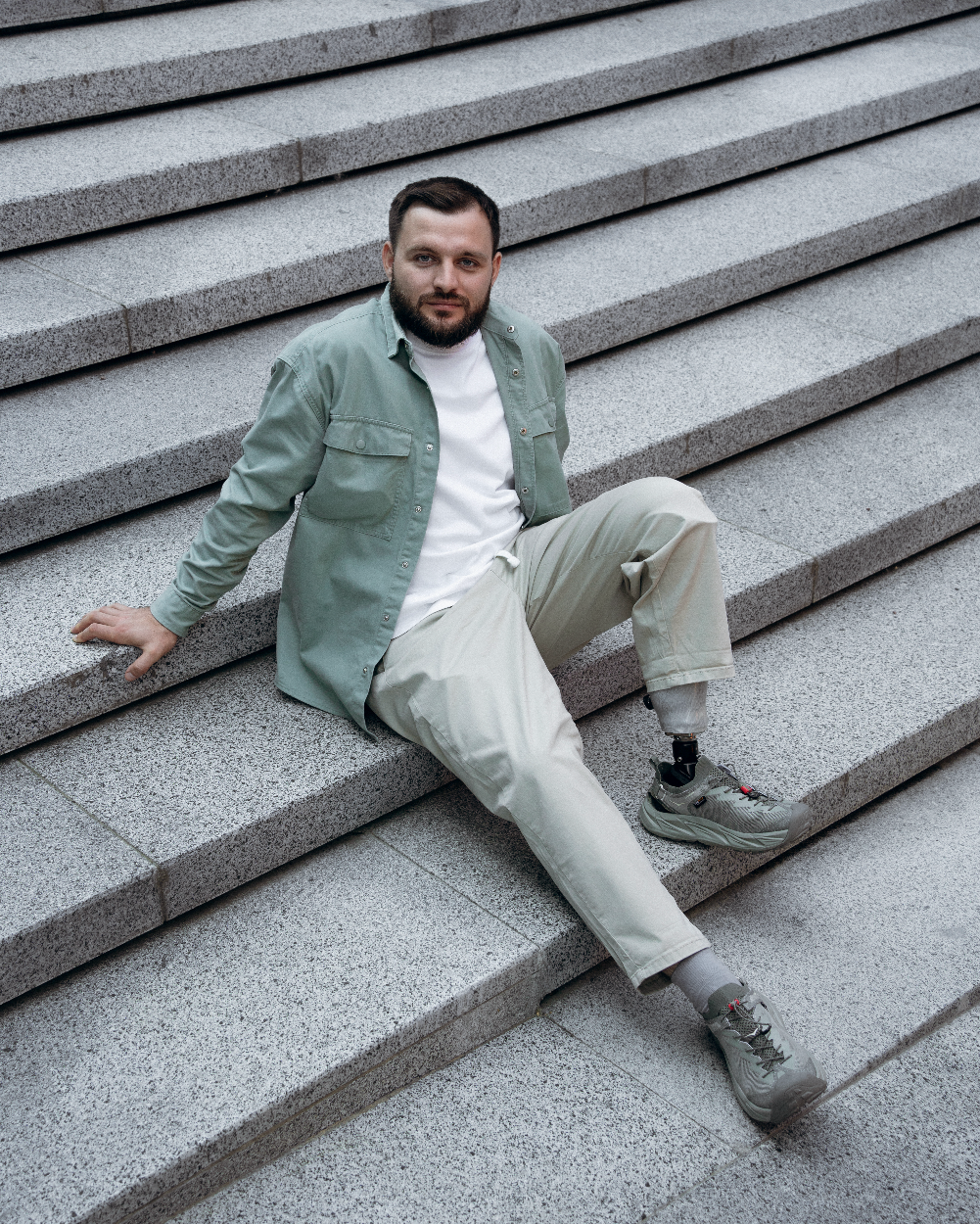 Man with a prosthetic foot sitting on concrete steps wearing a light green jacket and beige pants.
