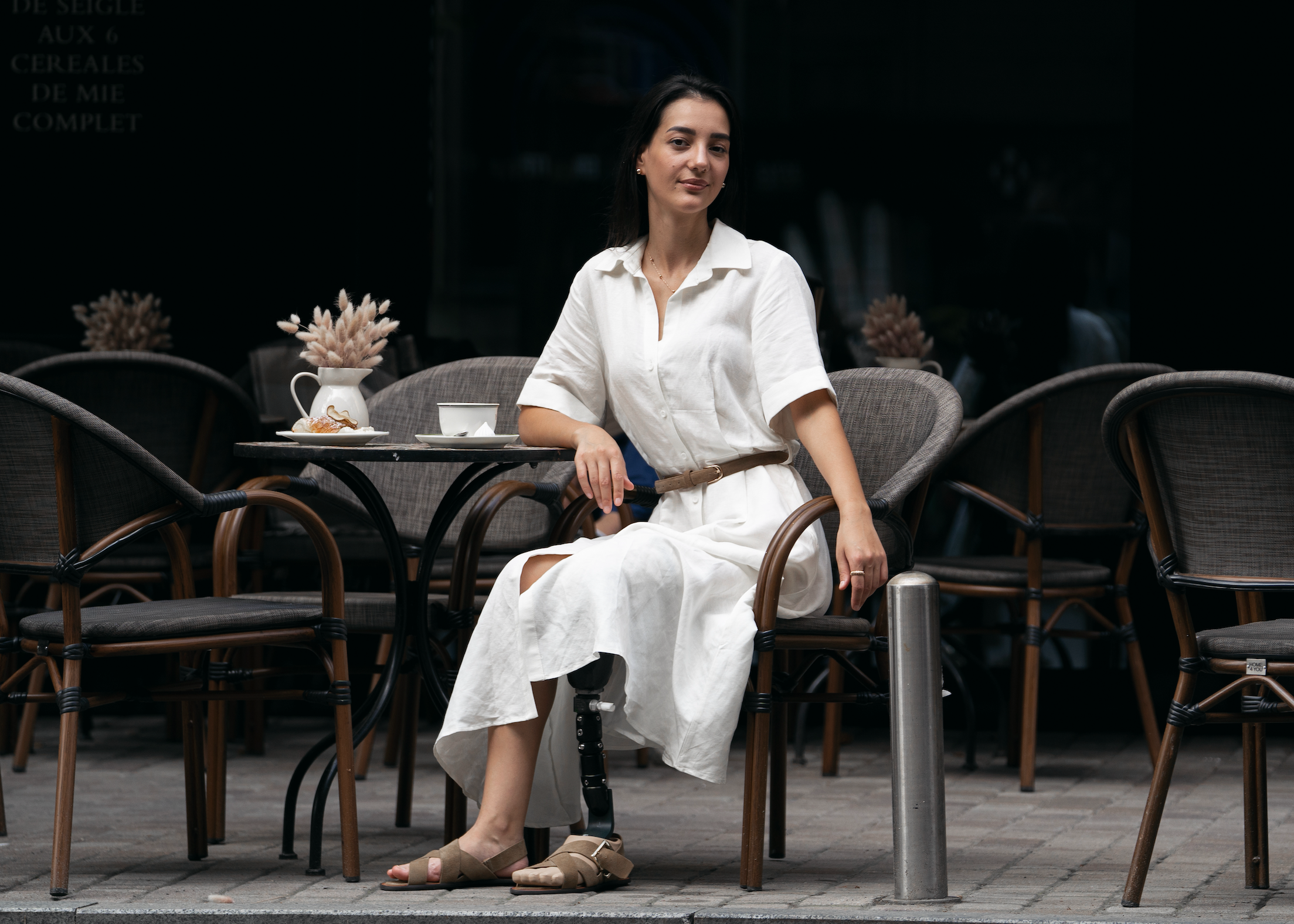 A woman sits at an outdoor cafe table wearing Forever S prosthetic foot in a white short-sleeve button-up dress with a brown belt. The setting features dark wicker chairs and a dark cafe facade in the background.